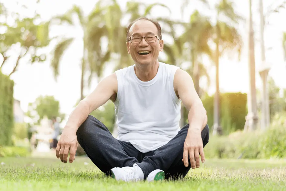 Older man in exercise attire sitting on the grass with a big smile.